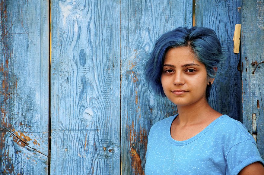 Young woman with blue hair stands in front of a blue fence wearing a blue shirt.